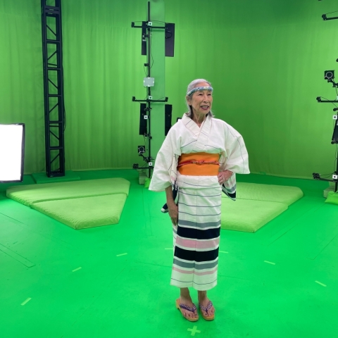 a woman dressed in Japanese kimono and headband stands in a green room filled with lights and cameras